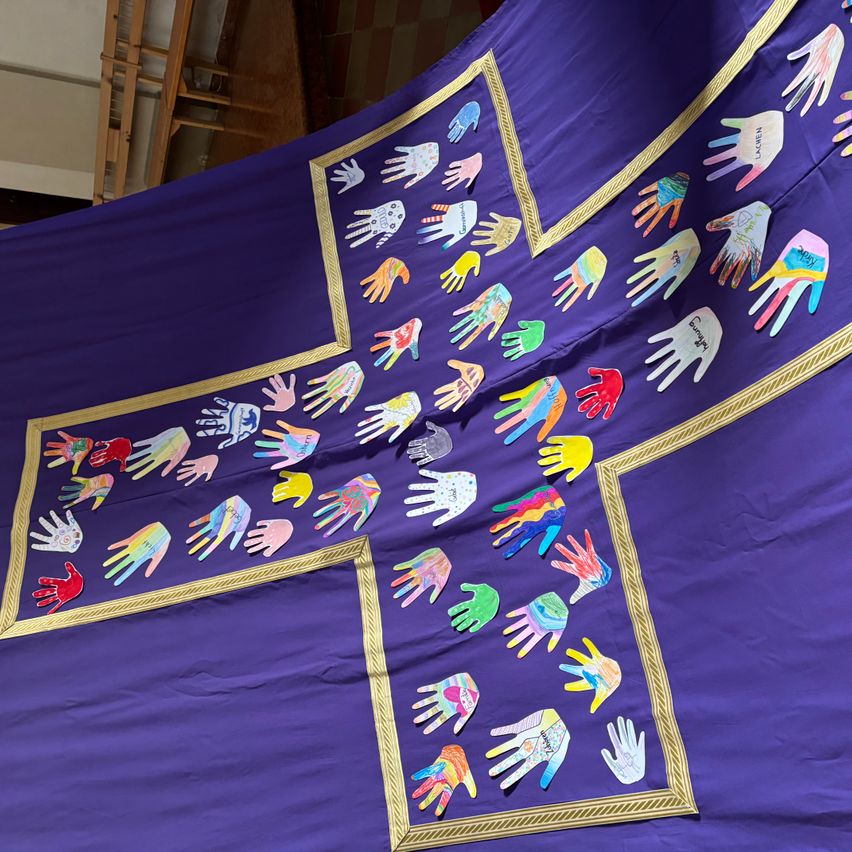A large purple banner with a cross design, featuring numerous handprints in various colors. The banner has golden edges and a golden cross design.