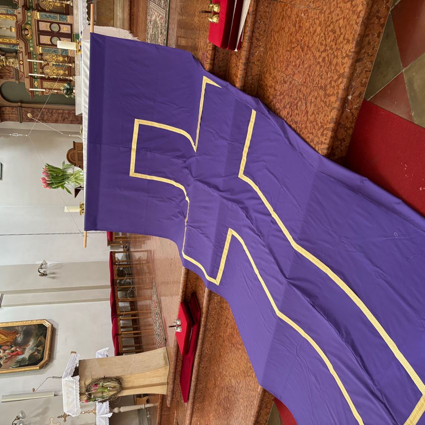 A purple cloth with a yellow cross design is draped over a wooden altar. The background shows a church interior with chairs and a painting.