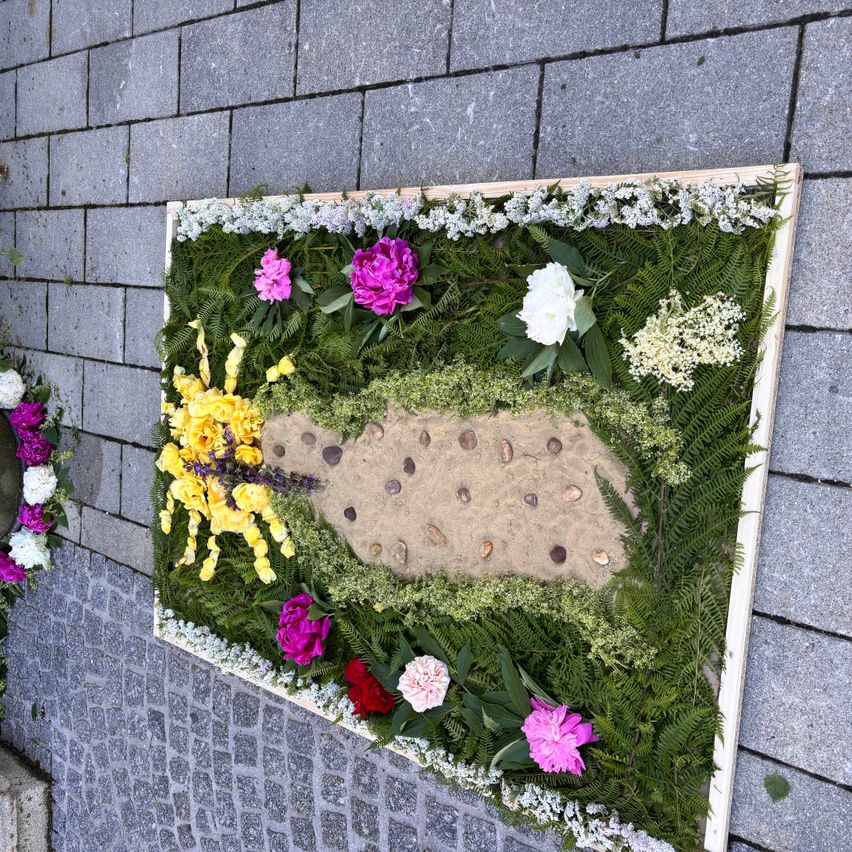 A rectangular arrangement of flowers and foliage on a brick wall. The design features a sand path with seashells, surrounded by a variety of flowers.