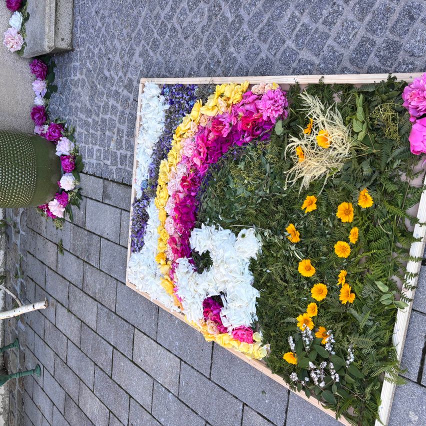 A rectangular wooden box filled with colorful flowers and green leaves. The flowers form a spiral design with various colors. A part of the wall is visible in the background.