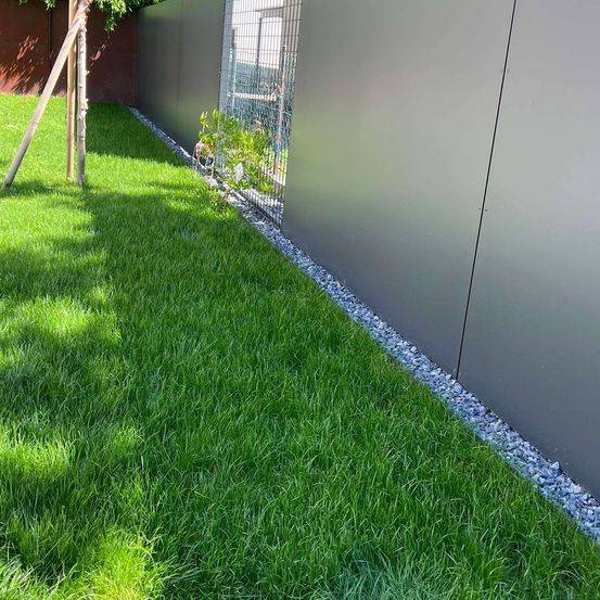 Ein Hinterhof mit einem sauber geschnittenen Rasen und einem grauen Kiesrand, neben einer grauen Wand mit einem Metallzaun.