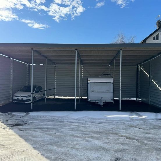 Ein weißer Wagen und ein weißer Anhänger sind unter einem Metallcarport in einem verschneiten Bereich geparkt. Der Carport hat ein Metalldach und Wände.