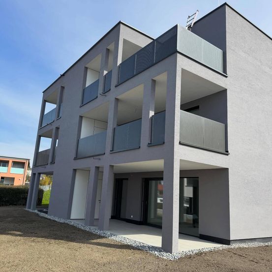 Ein modernes Apartmentgebäude mit grauer Außenfassade, mehreren Balkonen und einem Glaseingang. Das Gebäude hat ein Flachdach und ist von Kies umgeben.