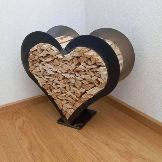 Ein herzförmiger Metallständer, gefüllt mit Holzstücken, steht auf einem Holzboden an einer weißen Wand.