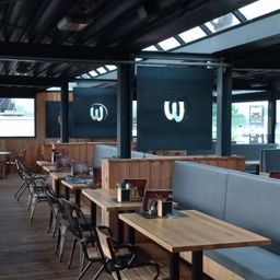 Ein leeres Restaurant mit hölzernen Sitzbänken und Tischen. Ein großes W-Logo hängt an der Decke. Das Restaurant hat große Fenster und einen Holzboden.