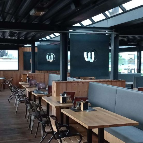Ein leeres Restaurant mit hölzernen Sitzbänken und Tischen. Ein großes W-Logo hängt an der Decke. Das Restaurant hat große Fenster und einen Holzboden.