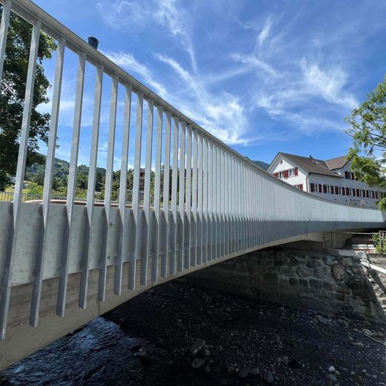 Eine moderne Brücke mit schlankem Design überspannt einen Fluss mit felsigen Ufern und einem klaren blauen Himmel im Hintergrund.
