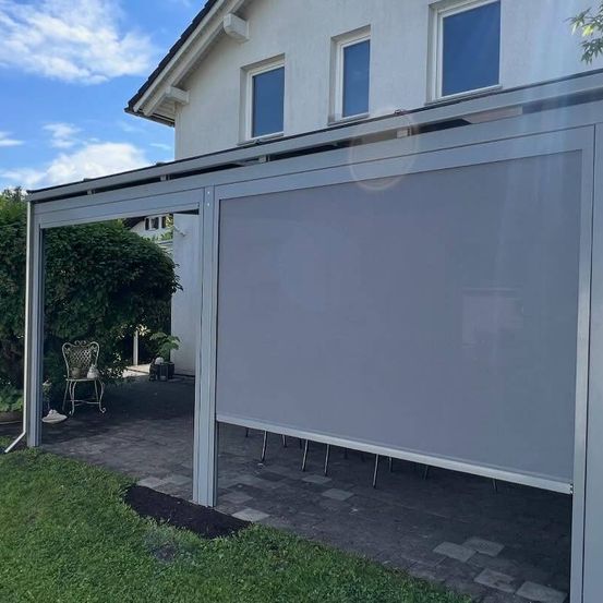 Ein grauer Rollladen erstreckt sich von einem weißen Gebäude mit Glasfenstern. Eine leere Stuhl und Pflanzen sind in der Nähe. Der Boden ist mit Steinen gepflastert.