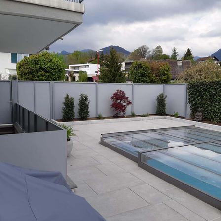 Ein Hinterhof mit Terrasse und Pool, umgeben von einem weißen Zaun. Bäume, Pflanzen und Büsche sind am Boden. Ein Gebäude ist in der Ferne, und Berge sind am Horizont.
