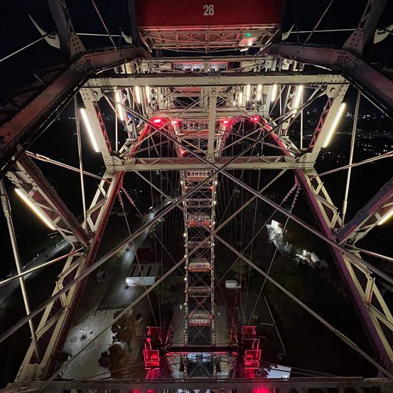 Ein Riesenrad bei Nacht mit roten Lichtern und der Nummer 28 sichtbar auf der Spitze. Die Struktur ist aus Metall mit mehreren Balken und Seilen.