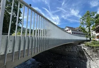 Eine moderne, weiße Brücke überspannt einen Fluss mit Bäumen und Häusern im Hintergrund unter einem klaren blauen Himmel.