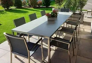 Ein Esstisch im Freien mit sechs Stühlen und einem Blumentopf darauf steht auf einer Terrasse mit grünem Rasen und Pflanzen.