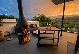 Eine Dachterrasse mit Kamin, Sofa, Tisch und Rollwagen. Die Terrasse bietet einen Blick auf einen Sonnenuntergang über einem Tal mit Bergen.