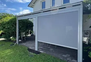 Eine weiße, einziehbare Überdachung befindet sich in einem Garten, umgeben von grünem Rasen und Pflanzen. Dahinter steht ein weißes Haus mit Fenstern und einer Terrasse.