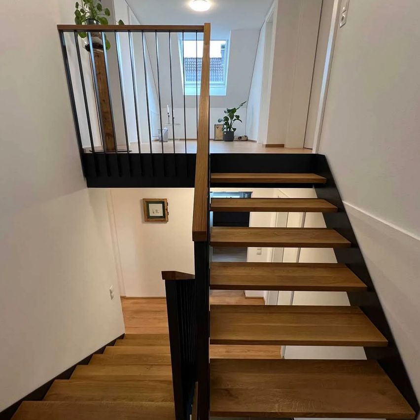 Eine moderne Treppe mit Holzstufen und schwarzen Geländern. Ein Topf mit Pflanzen befindet sich auf dem Balkon im zweiten Stock.