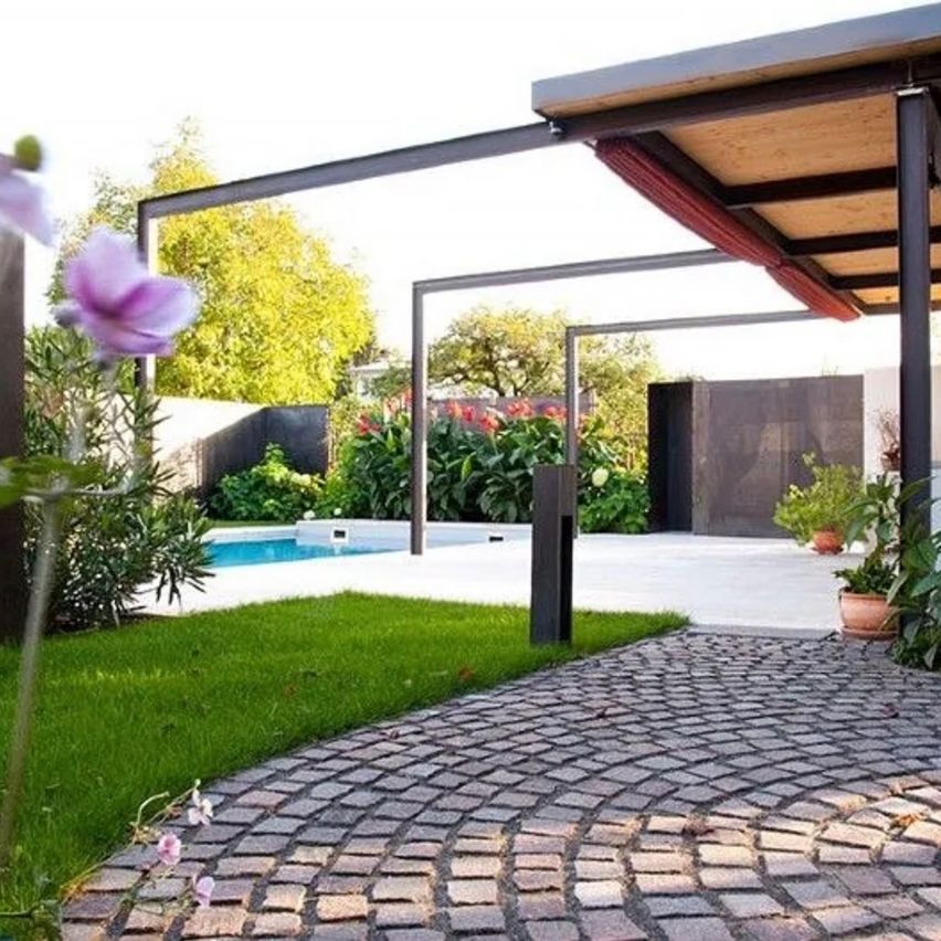 Ein Hinterhof mit einem gepflasterten Weg, der zu einem Pool führt. Eine Terrasse mit Metallgeländern und einem Holzdach. Der Bereich ist von Pflanzen und Bäumen umgeben.