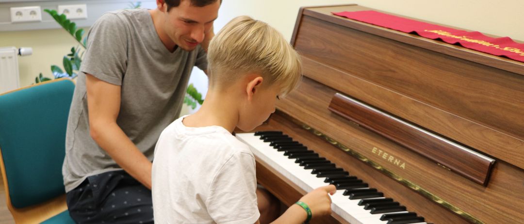 Ein junger Junge spielt Klavier, während ein Mann zuschaut, beide sitzen drinnen mit einem Fenster im Hintergrund.
