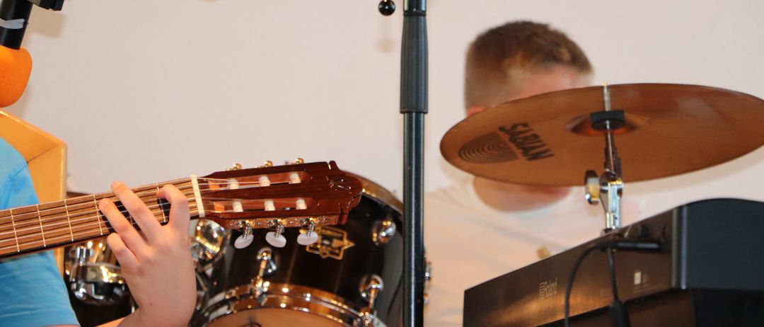 Ein Musiker spielt Gitarre, während ein anderer Schlagzeug spielt. Ein Becken und eine Tastatur sind ebenfalls zu sehen.