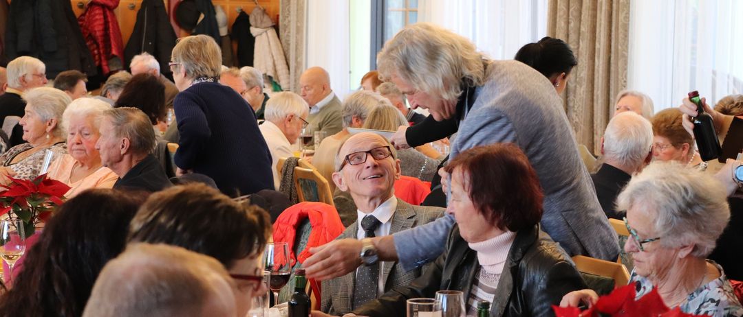 Eine Gruppe älterer Erwachsener versammelt sich in einem Raum, einige sitzen an Tischen, andere stehen. Eine ältere Frau hilft einem älteren Mann mit Brille. Ein Tisch im Vordergrund hat Weingläser, Flaschen und eine rote Blumenarrangement.