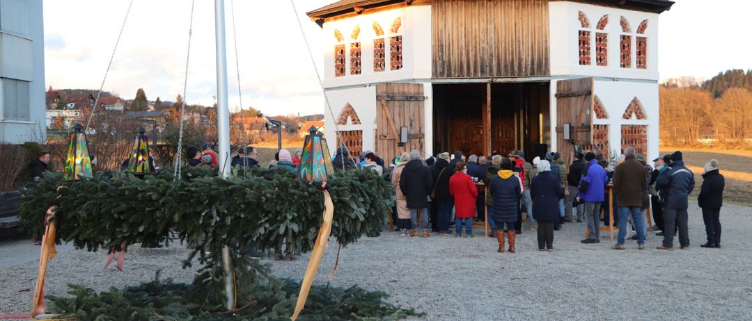 Eine Gruppe von Menschen steht vor einer Kirche mit einem Weihnachtskranz, der an einem Pfahl hängt. Die Kirche hat bogenförmige Fenster und eine große offene Tür.