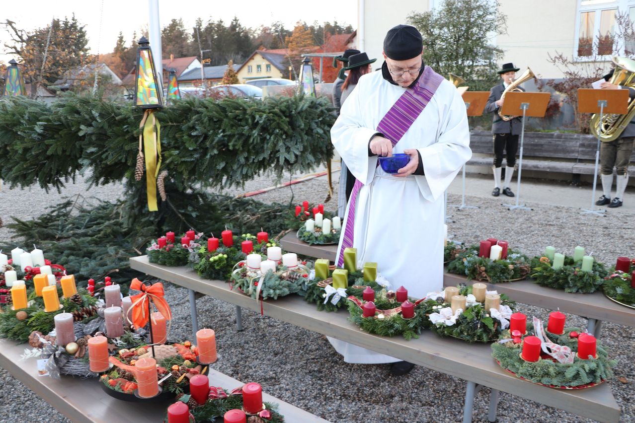 Ein Priester steht vor einem Tisch mit Kränzen und Kerzen und hält eine blaue Tasse. Hinter ihm spielt eine Person ein Blechinstrument. Bäume und Gebäude sind im Hintergrund.
