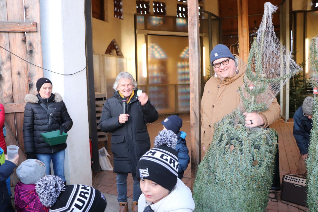 Ein Mann mit einem Mikrofon steht neben einem Weihnachtsbaum und lächelt. Ein Junge in einer weißen Jacke und einem blauen Hut schaut zu. Eine weitere Person in einem schwarzen Mantel hält eine grüne Box. Im Hintergrund befindet sich ein Gebäude mit einer Glastür.