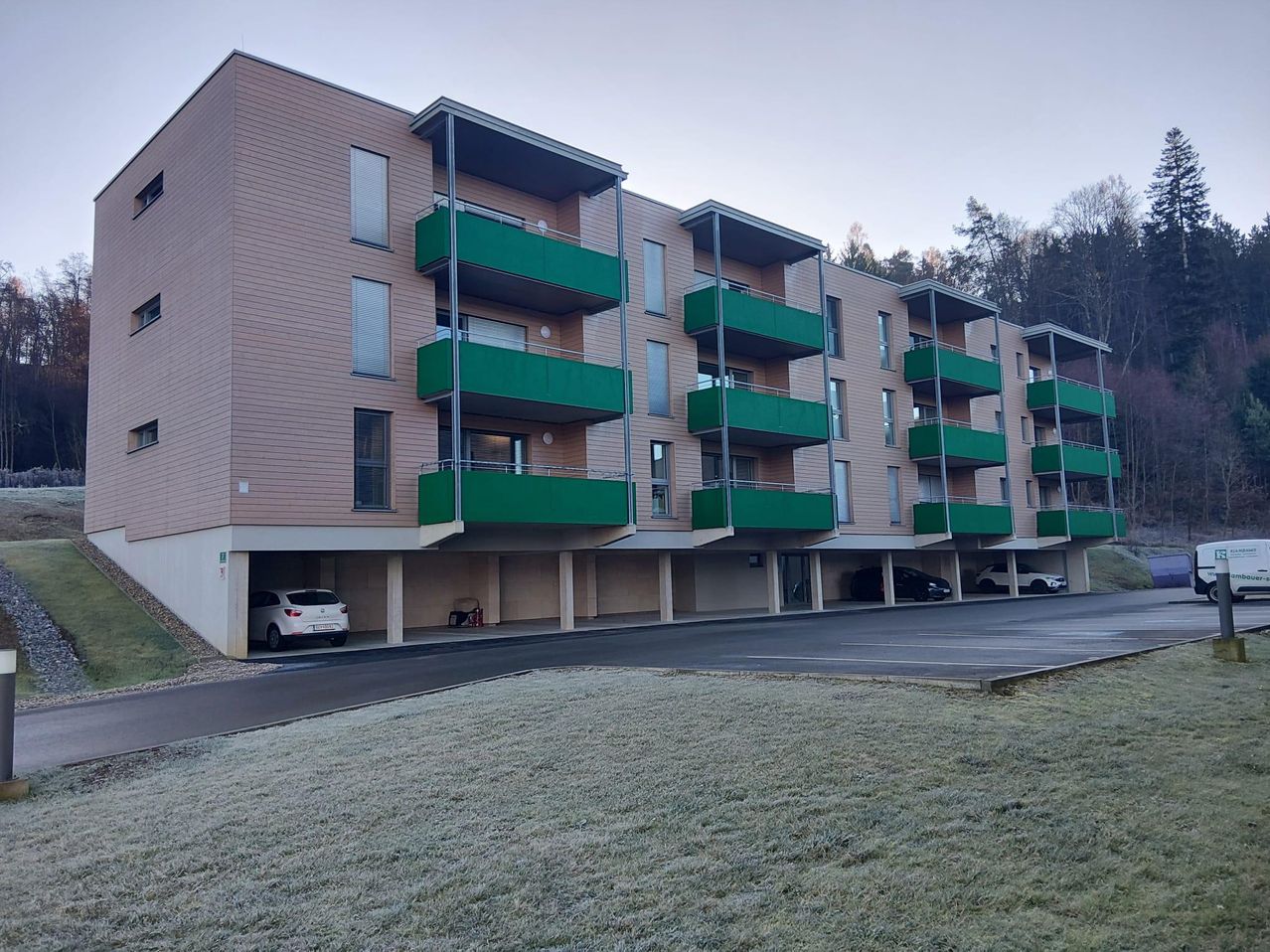 Ein mehrstöckiges Apartmentgebäude mit beigefarbener Außenfassade und grünen Balkonen. Vor dem Gebäude sind mehrere Autos geparkt.