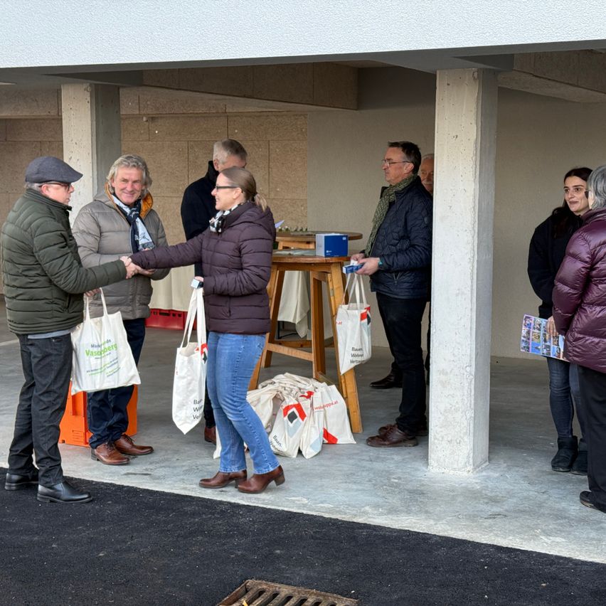 Eine Gruppe von Menschen steht in einem überdachten Bereich, vielleicht einer Garage. Sie halten weiße Taschen und einige schütteln Hände. Ein Tisch mit einer Aufbewahrungsbox und Taschen steht vor ihnen.