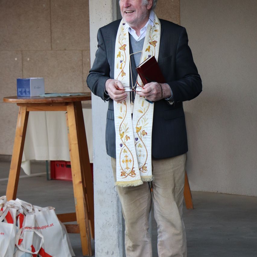 Ein älterer Mann mit einer weißen Stola steht neben einer Säule, hält ein Buch und eine Brille in der Hand. Hinter ihm steht ein Holztisch mit einer Schachtel. Taschen liegen auf dem Boden.