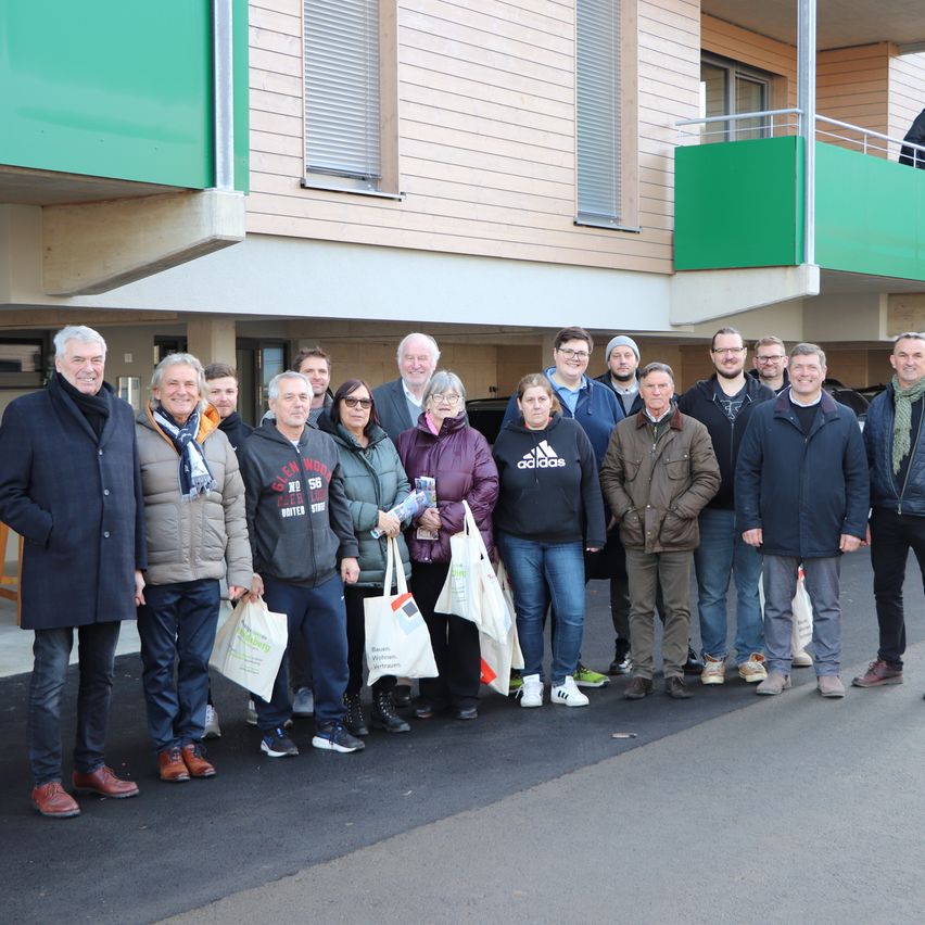 Eine Gruppe von Menschen in Winterkleidung steht vor einem modernen Gebäude mit grünen Akzenten. Sie halten Einkaufstüten und lächeln für ein Foto.