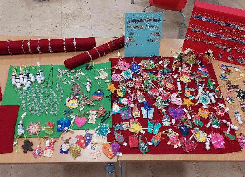 Ein Tisch zeigt verschiedene farbenfrohe handgefertigte Weihnachtsornamente und Schmuck. Links auf einem grünen Tuch befinden sich weiße Kugeln und Schneeflocken. In der Mitte hat ein rotes Tuch viele Ornamente mit verschiedenen Designs. Rechts halten blaue und rote Tücher Ohrringe.