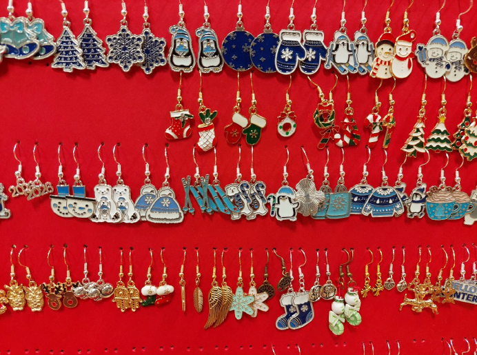 Eine Sammlung von weihnachtlich gestalteten Ohrringen wird auf rotem Hintergrund präsentiert. Die Ohrringe zeigen verschiedene Designs wie Schneemänner, Weihnachtsbäume und Wintermotive. Jeder Ohrring ist einzigartig gestaltet, einige mit Glitzer oder Emaillefarben. Die Ohrringe hängen an einem roten Pegboard, möglicherweise zum Verkauf.