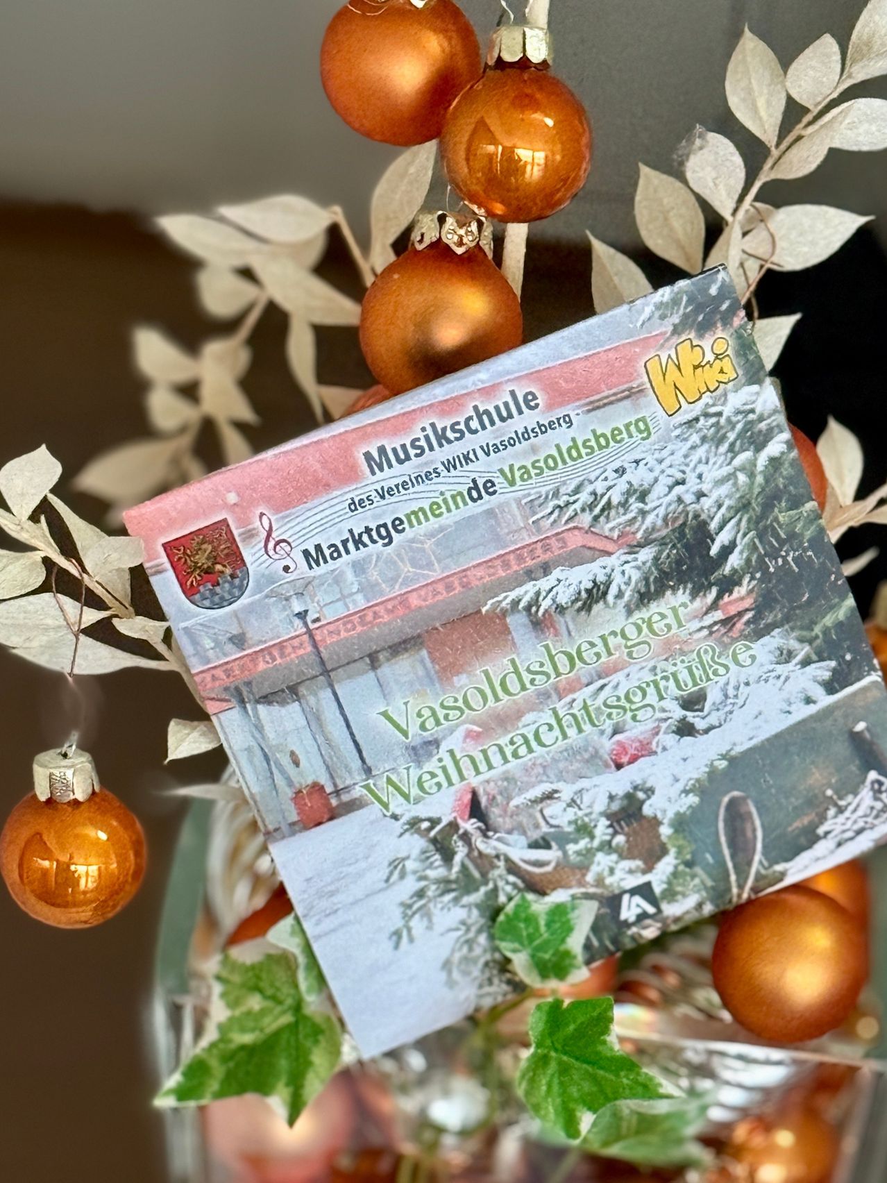 Eine Weihnachtskarte mit einer verschneiten Szene eines Hauses. Es steht 'Marktge meinde Vasoldsberg' und 'Vasolsberger Weihnachtsgrüsse'. Sie befindet sich zwischen dekorativen Blättern und Ornamenten.