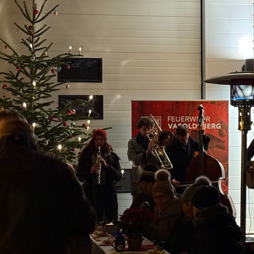 Eine kleine Band spielt Instrumente in einem Raum mit einem geschmückten Weihnachtsbaum, wo sich Menschen versammelt haben. Ein Banner mit dem Text 'Feuerwir Vasoldberg' ist sichtbar.