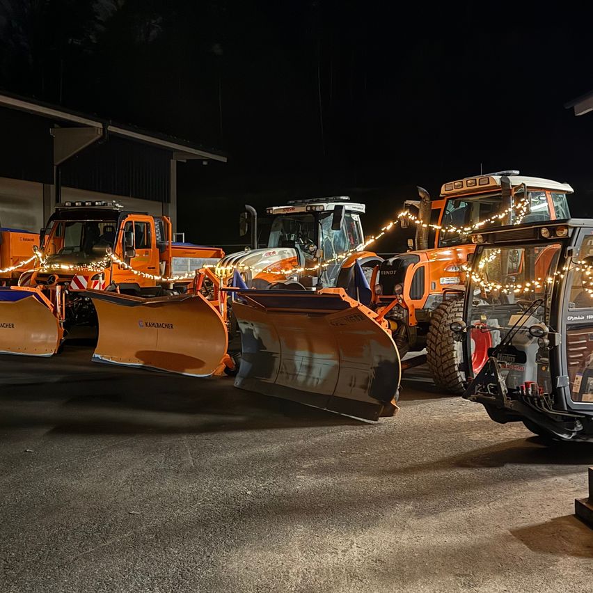 Vier orangefarbene Schneepflüge sind in einer Reihe geparkt, geschmückt mit Lichtern und Flaggen. Sie sind nachts in einem Parkplatz beleuchtet.