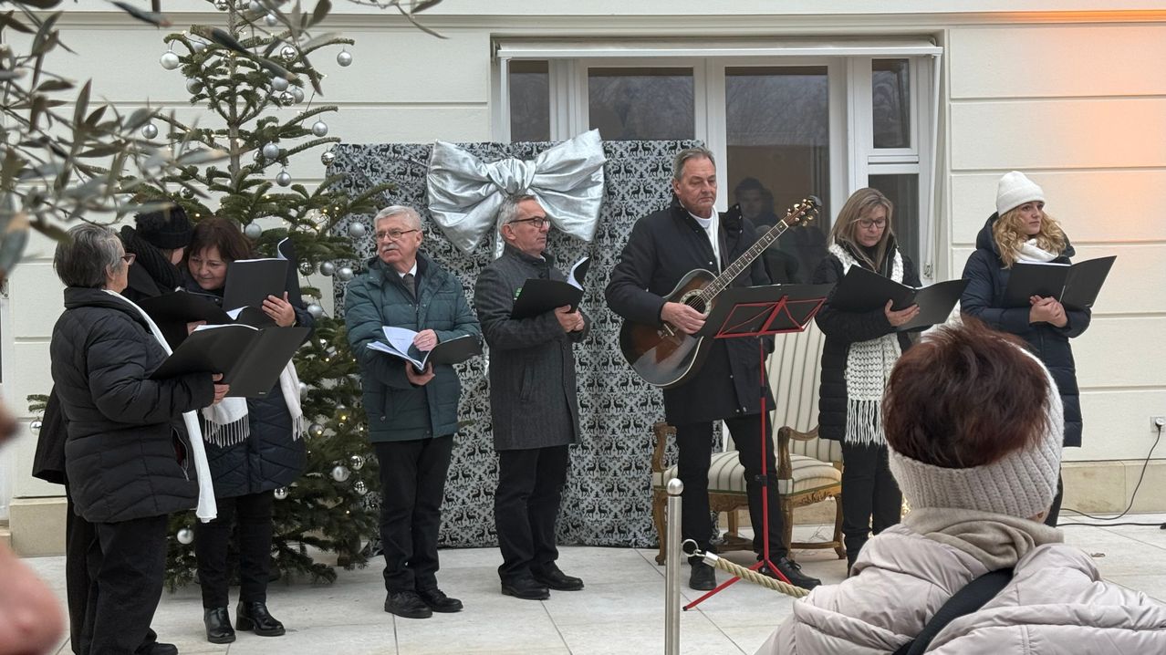 Vier Personen, darunter ein Mann mit Gitarre, stehen vor einem Weihnachtsbaum und lesen Notenblätter. Dahinter befindet sich eine dekorierte Wand mit einer silbernen Schleife.