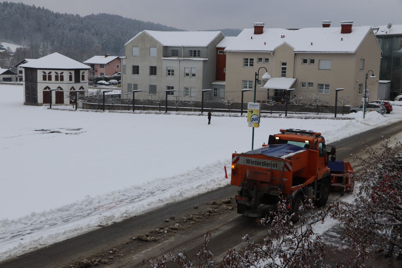 Ein Schneepflug fährt auf einer verschneiten Straße vor Häusern. Es gibt ein Schild am Bürgersteig und eine Person geht auf dem Schnee.