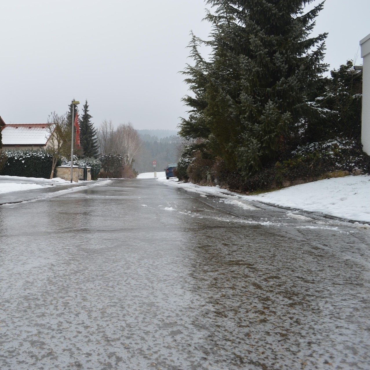 Eine verschneite Straße mit Bäumen und Büschen, die mit Schnee bedeckt sind. Ein Auto ist am Ende der Straße geparkt.