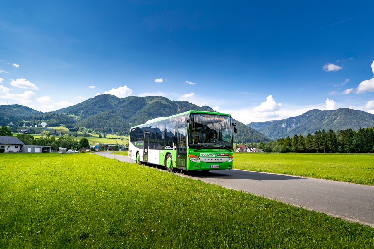 Ein grüner und weißer Bus fährt eine Bergstraße hinunter, mit Bergen und blauem Himmel im Hintergrund.