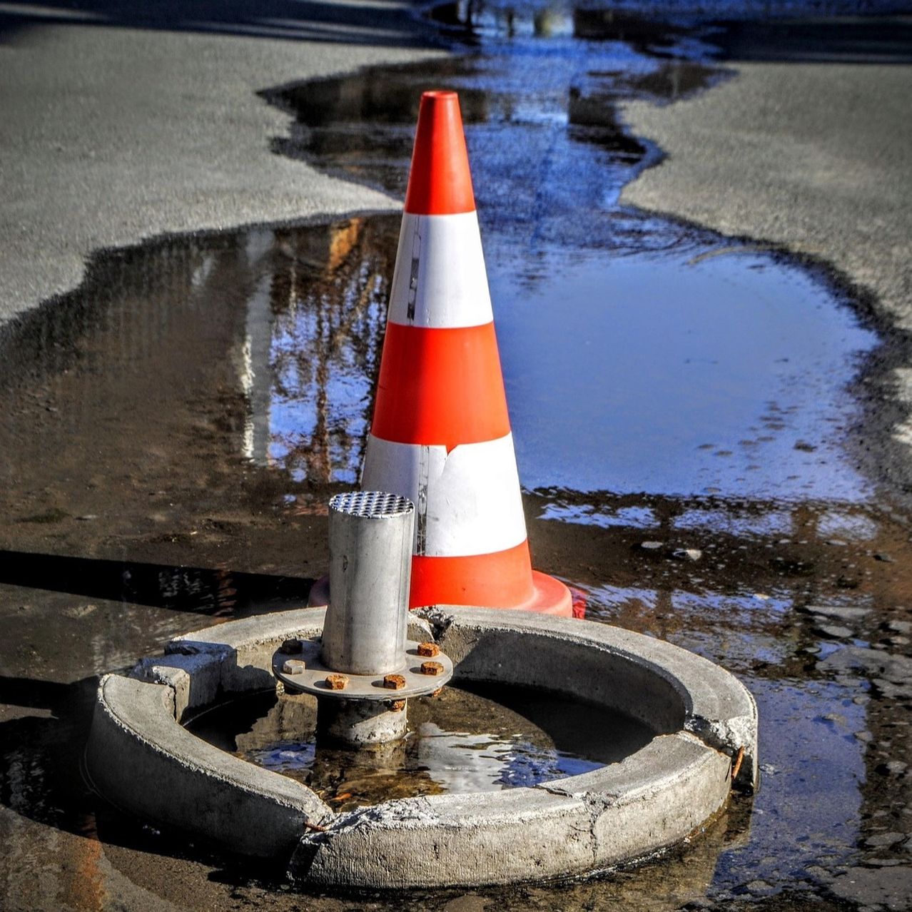 Ein mit Wasser gefülltes Schlagloch ist auf einer Straße sichtbar, wobei ein Verkehrskegel daneben steht. Der Kegel ist orange und weiß und teilweise im Wasser eingetaucht.