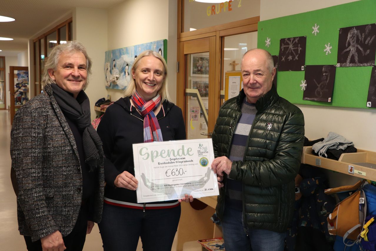 Drei Menschen stehen lächelnd in einem Raum. Die Frau in der Mitte hält ein Zertifikat mit der Aufschrift 'Spende' und '630'. Der Mann rechts trägt eine grüne Jacke und der Mann links eine graue Jacke. Hinter ihnen befindet sich eine Wand mit einem grünen Banner und einer Holztür.