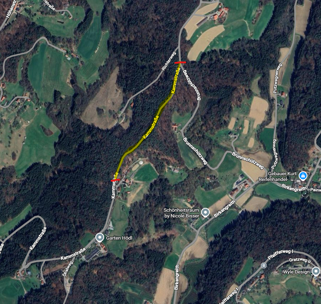 Luftaufnahme eines ländlichen Gebiets mit grünen Feldern, Wäldern und einer kurvenreichen Straße, die mit einer gelben Linie markiert ist. Ein roter Pfeil zeigt auf einen bestimmten Bereich. Das Bild zeigt auch Gebäude und einen Golfplatz.