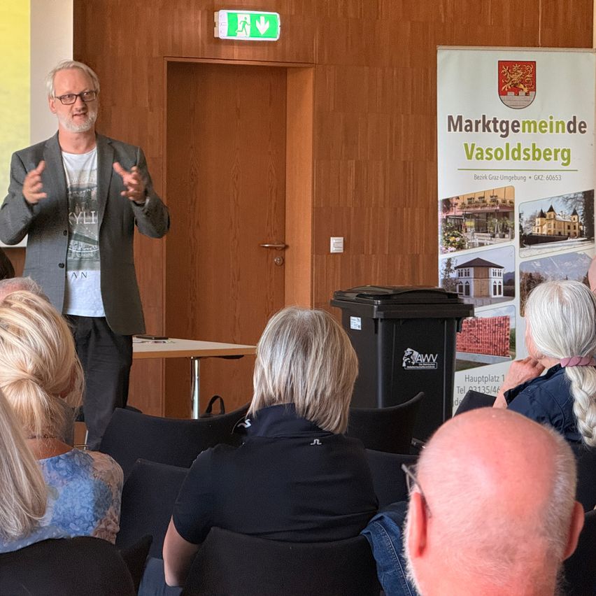 Ein Mann hält eine Präsentation vor einem Publikum, steht vor einer Holzwand mit einem Notfallausgangsschild und einer Werbung für Marktgemeinde Vasoldsberg.