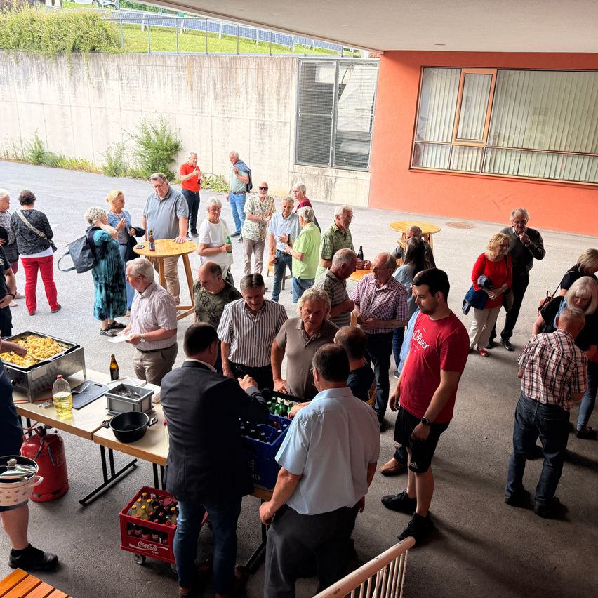 Eine Gruppe von Menschen versammelt sich im Freien für eine Veranstaltung, mit Tischen, Essen und Getränken. Einige tragen Freizeitkleidung, andere formelle Kleidung. Ein Gebäude mit Glasfenstern steht hinter ihnen.