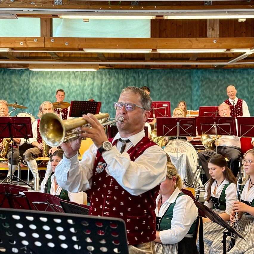 Ein Mann spielt Trompete in einem Raum mit einer Band. Der Raum hat Holzbalken und einen blauen Vorhang. Andere Bandmitglieder sitzen im Hintergrund und spielen verschiedene Instrumente.