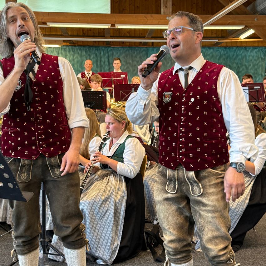 Zwei Männer stehen vor einer Gruppe, halten Mikrofone und singen. Hinter ihnen spielt eine Frau Klarinette, und eine Band ist sichtbar. Die Umgebung ist drinnen, mit sitzenden und stehenden Personen.