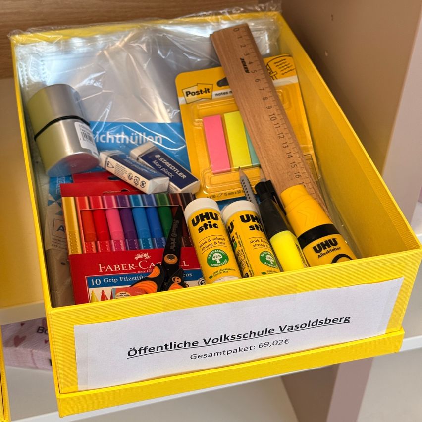 Eine gelbe Box enthält einen Lineal, Stifte, Kleber, Scheren und andere Schreibwaren. Die Box hat einen Aufkleber mit der Aufschrift 'Offentliche Volksschule Vasoldsberg Gesamtpaket: 69,02€'.