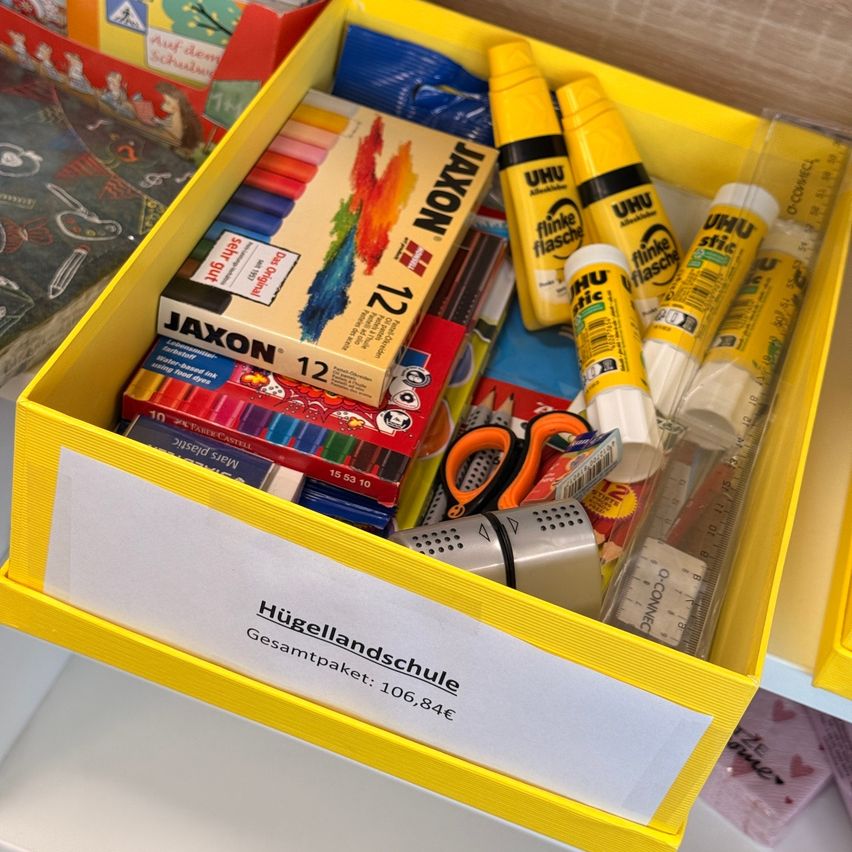Ein gelber Kasten mit der Aufschrift 'Hugellandschule' enthält Buntstifte, Klebestifte, Scheren und andere Schulmaterialien. Die Gesamtkosten betragen 106,84 Euro.