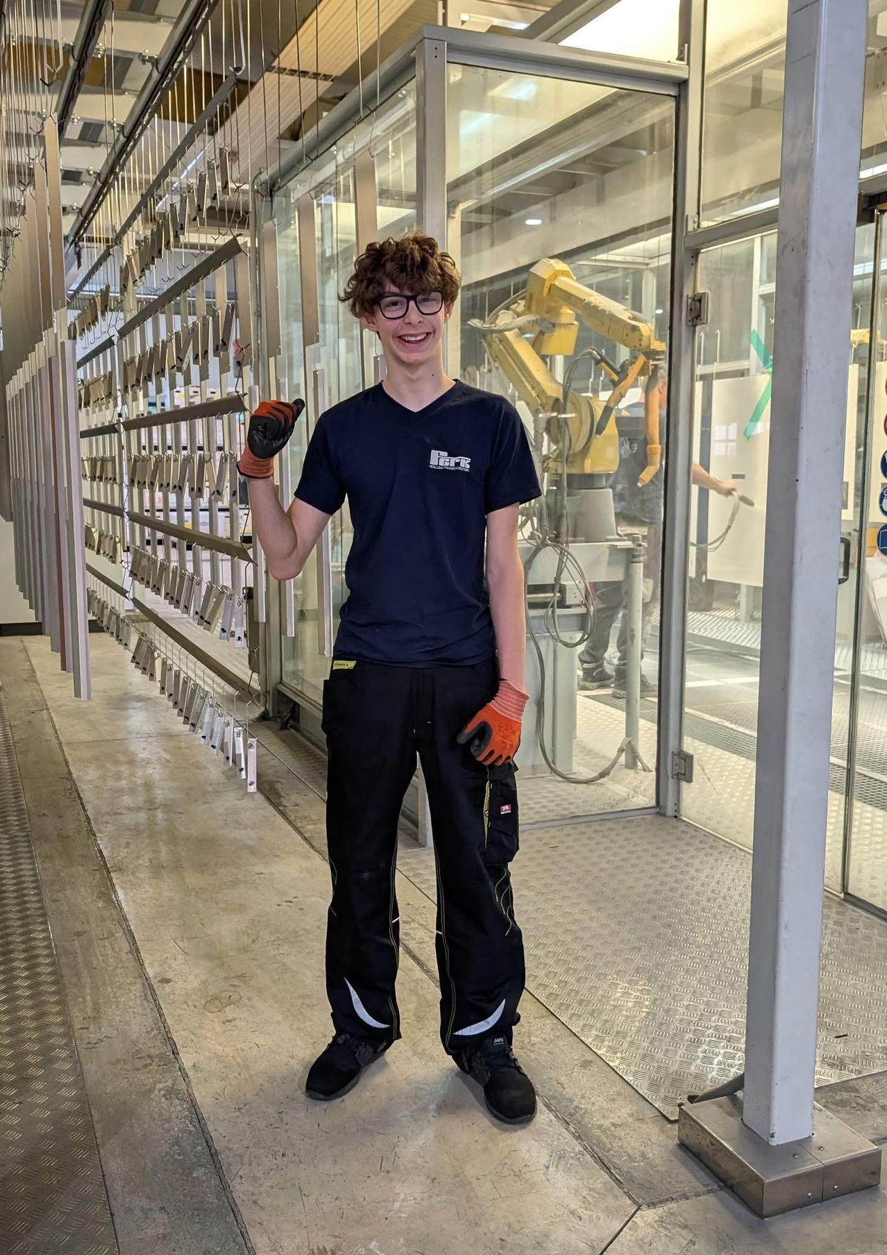 Ein junger Mann in einer Fabrikumgebung, der ein blaues T-Shirt und schwarze Hose trägt, lächelt für ein Foto, während er einen Handschuh hält. Hinter ihm ist eine Glaswand mit einem sichtbaren Roboterarm.