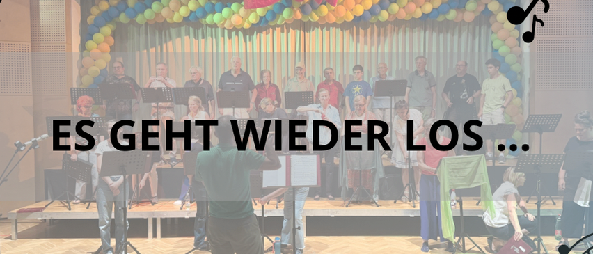 Ein Chor probet in einem Raum mit rotem Vorhang, Luftballons und Noten. Ein Mann steht vor ihnen und hält einen Notenständer.
